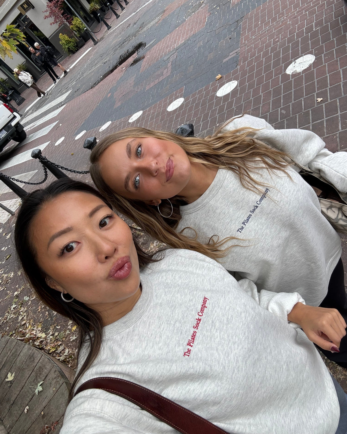 Two women taking a selfie outdoors on a brick sidewalk street corner, wearing matching light gray sweatshirts with 'The Pilates Sock Company' printed on the chest in red text.