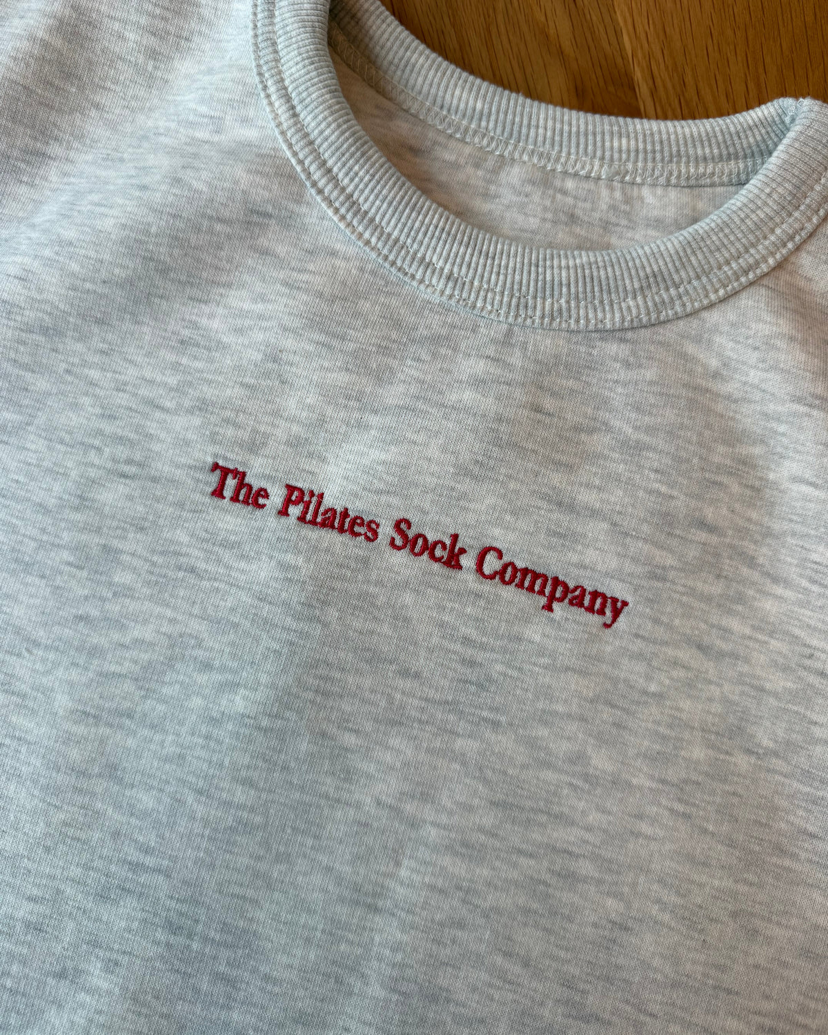 Close-up of a light heather gray crewneck sweatshirt laid flat on a wooden surface, featuring the text 'The Pilates Sock Company' embroidered in red across the chest.